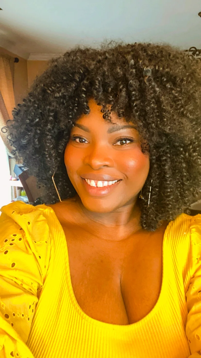 Kerri Ann Bryan smiles at the camera, wearing a bright yellow blouse and gold earrings, with voluminous curly dark hair framing her face.