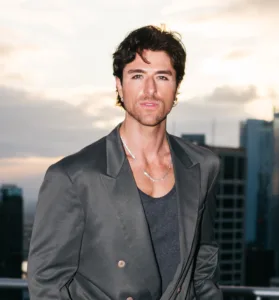 Anthony Randello-Jahn poses outdoors in a dark gray blazer and tank top with a city skyline blurred in the background.