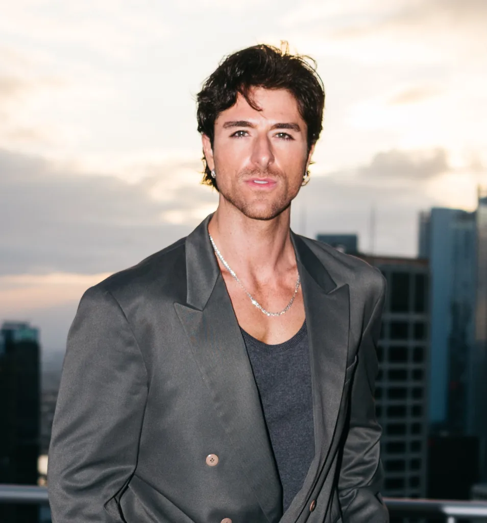 Anthony Randello-Jahn poses outdoors in a dark gray blazer and tank top with a city skyline blurred in the background.
