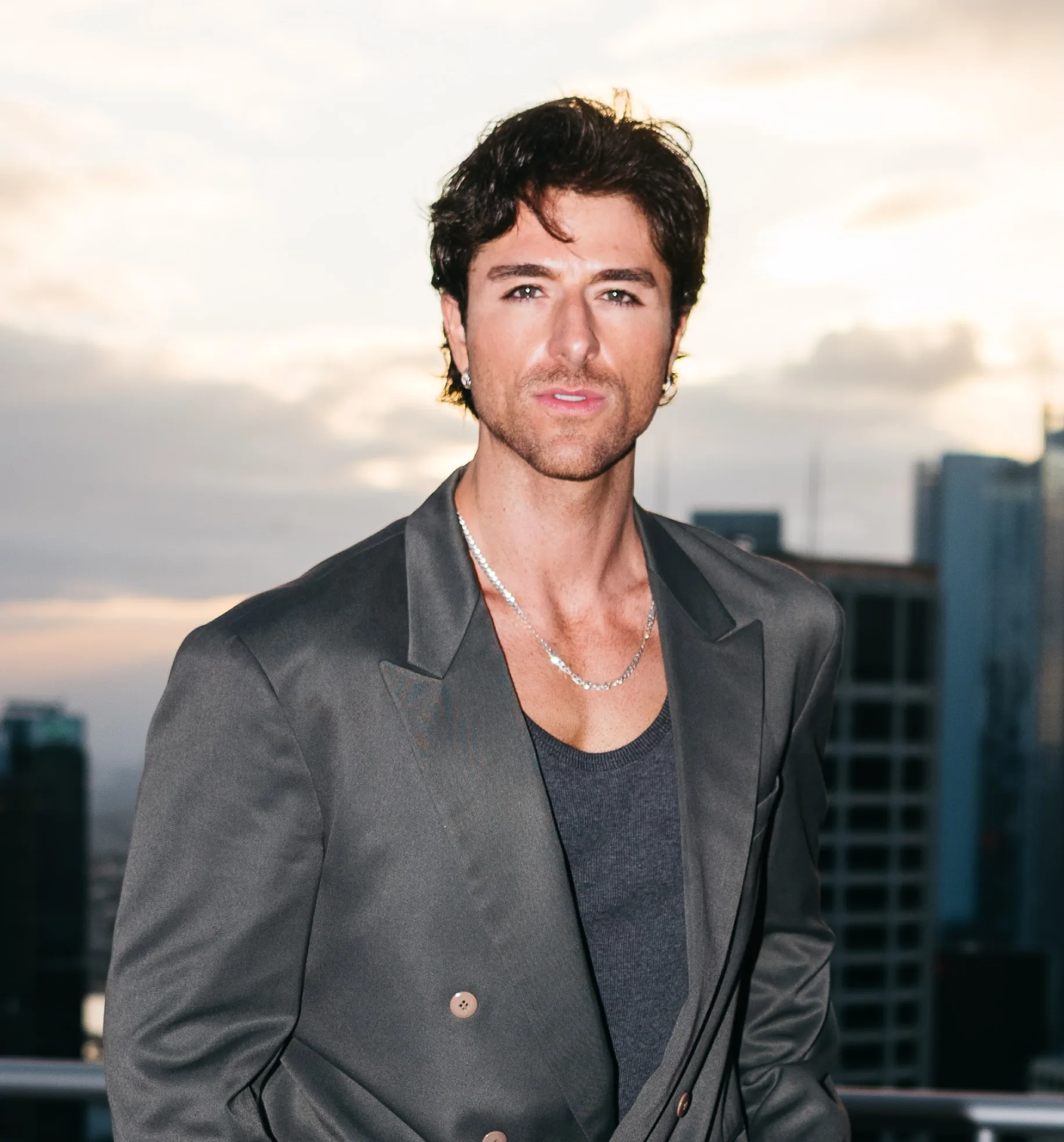 Anthony Randello-Jahn poses outdoors in a dark gray blazer and tank top with a city skyline blurred in the background.