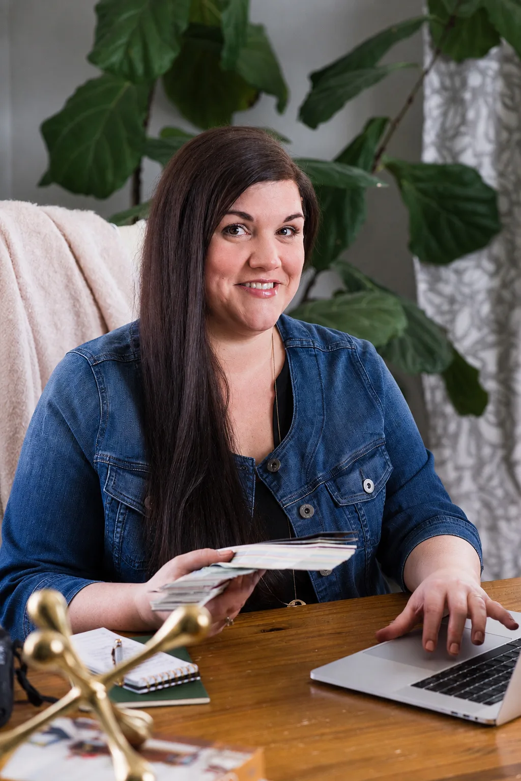 Cyd Converse sits at a desk holding a stack of color swatches while working on a laptop, with large leafy plants in the background.