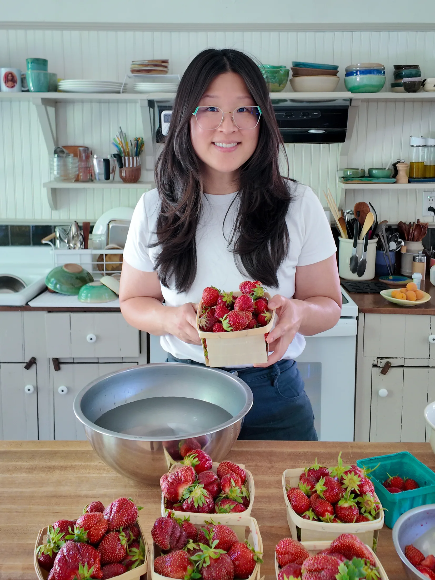 Jennifer Kim stands in a bright kitchen holding a basket of fresh strawberries, with more strawberries spread across the counter in front of her.