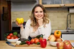 A woman smiles in her kitchen, leaning over her island with a countertop and sink behind her. She holds a yellow bell pepper and has other colorful fruits and veggies arranged on the island around her